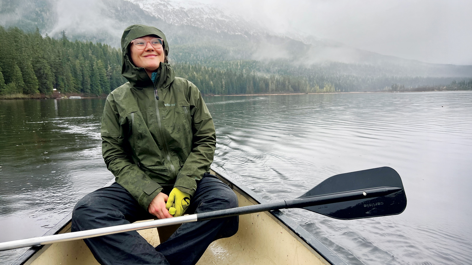 A person, embodying The Explorer, wears a green rain jacket and glasses while sitting in a canoe on a calm lake, surrounded by forest and misty mountains, holding a paddle. Inspired by adventurers like Tessa Hulls.