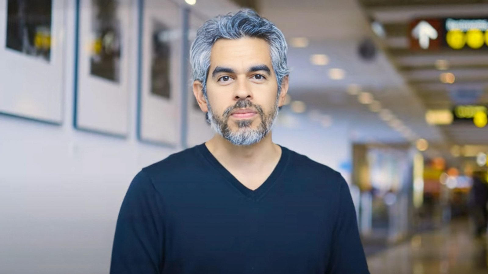 A man with gray hair and a beard, wearing a black V-neck shirt, stands indoors in a brightly lit hallway with signs and framed pictures—Civic Spacemaker Tommy Gregory is captured in this thoughtful moment.