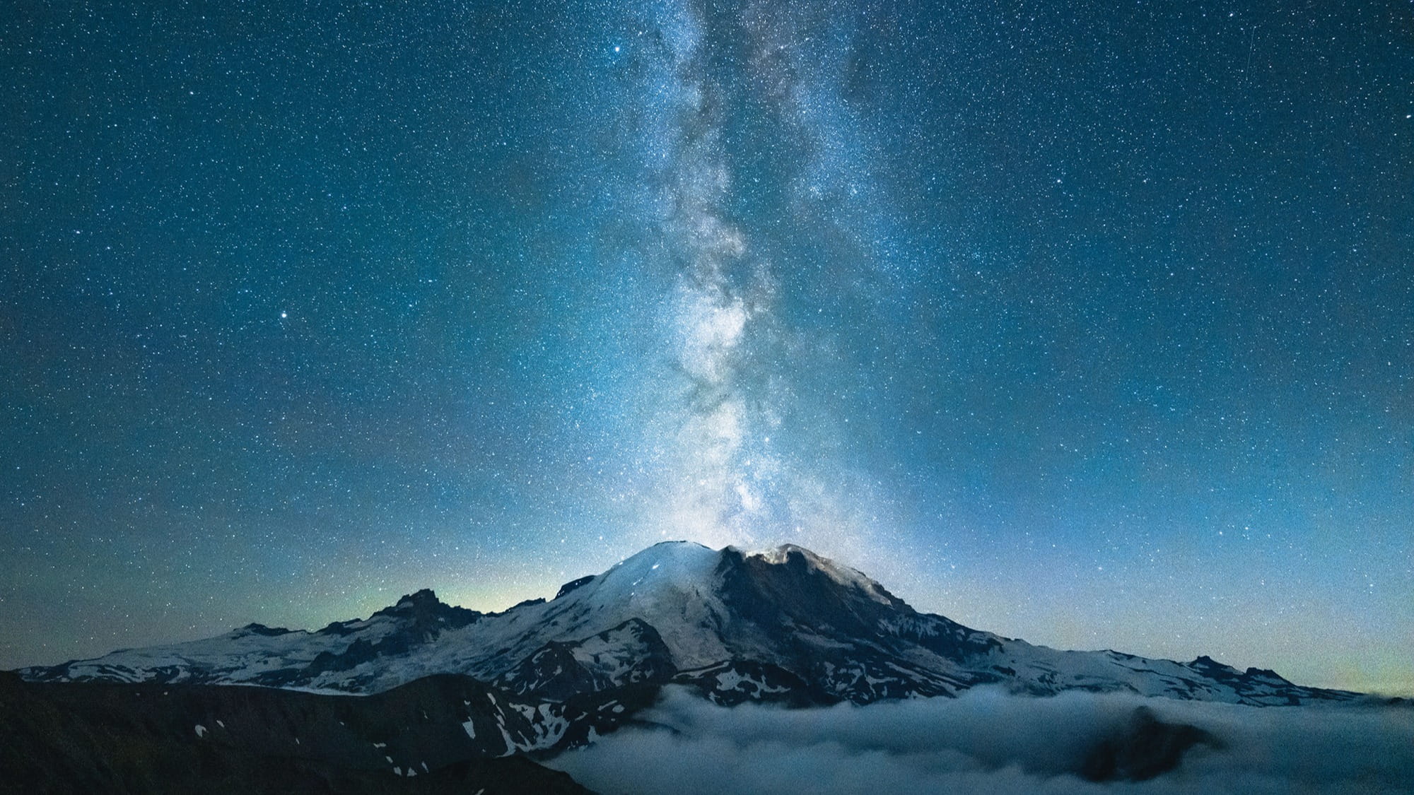 Snow-covered mountain under a clear night sky with the Milky Way galaxy visible above, perfect for hiking or photography. Captured by Tiffanie Yang, clouds and stars surround this breathtaking natural scene.
