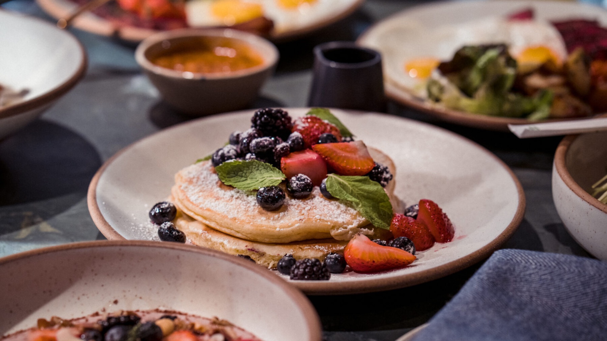A plate of pancakes topped with fresh strawberries, blackberries, blueberries, and mint leaves, surrounded by other breakfast dishes.