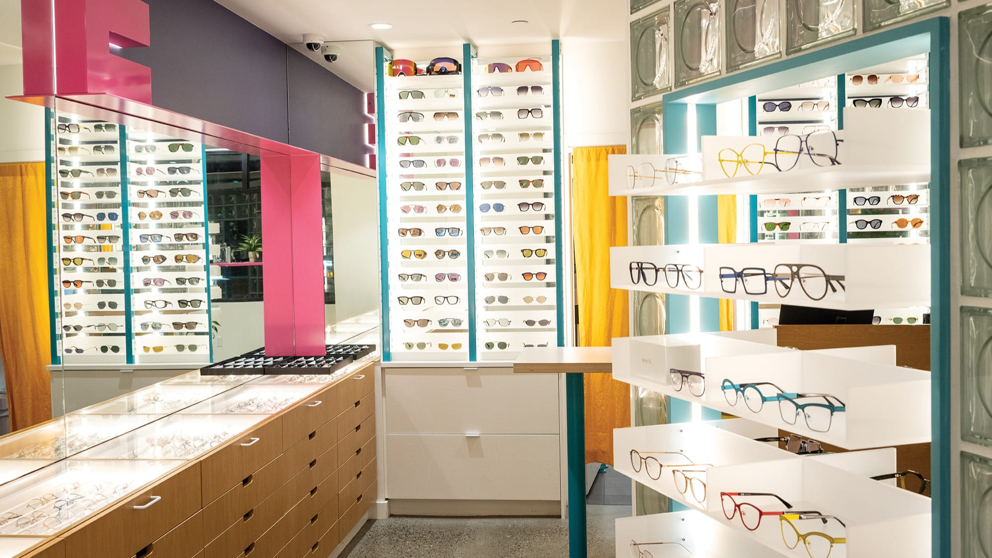 A brightly lit eyewear store, part of Dr. Will Pentecost’s optometry clinic Eye Eye, displays various eyeglasses and sunglasses on shelves and racks, with a counter and mirror in the foreground.