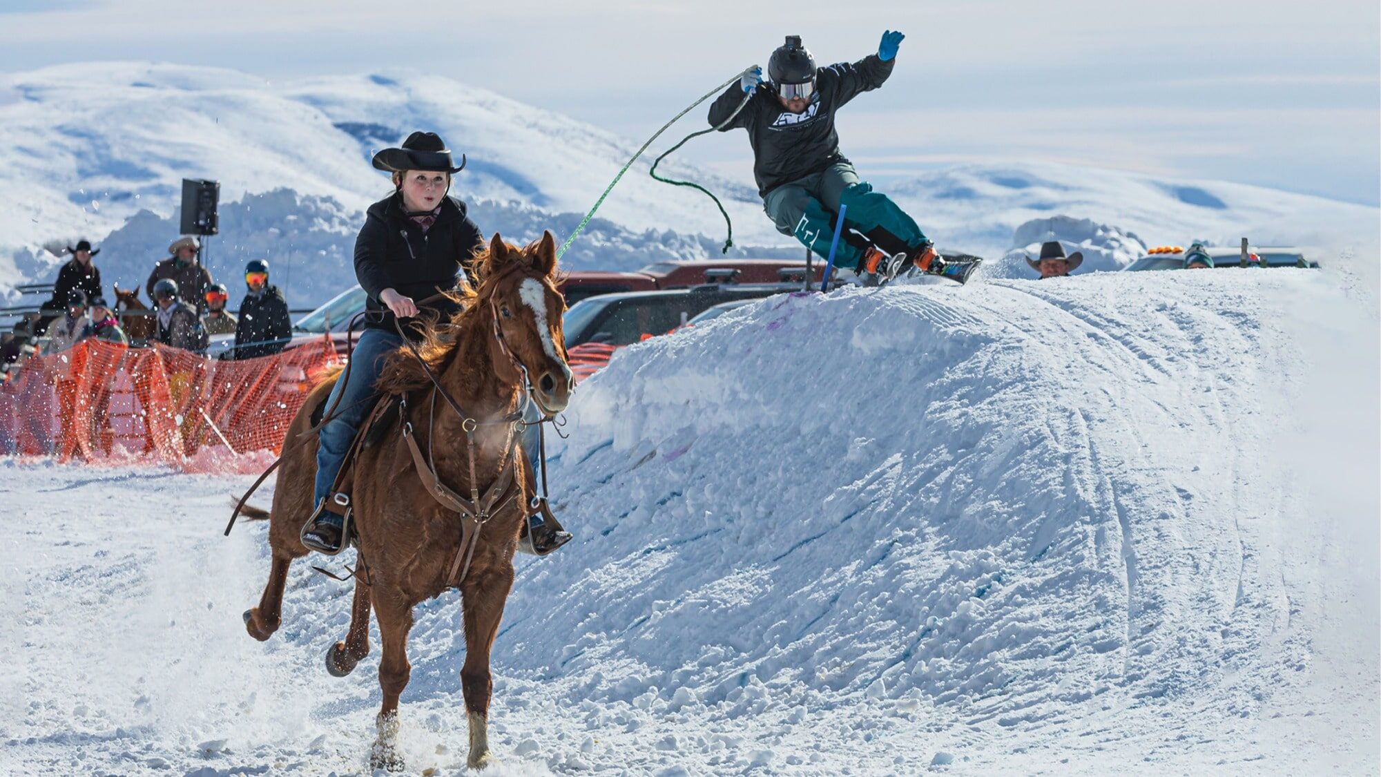 A person on horseback pulls a skier who jumps over a snow ramp during a skijoring event with spectators and mountains in the background.