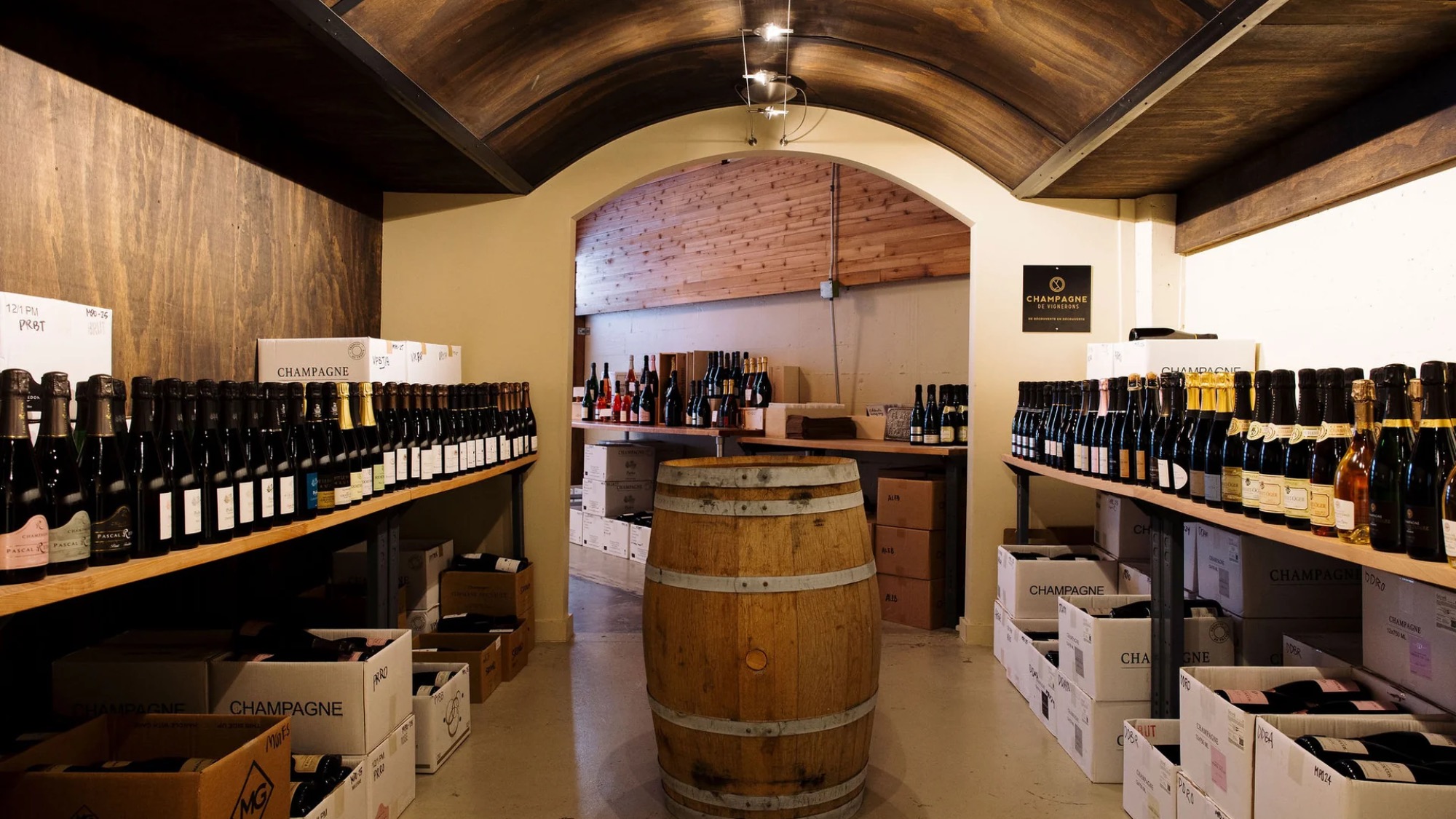 Wine bottles are displayed on wooden shelves along both walls of a cellar, with a barrel in the center and boxes of wine stacked on the floor.