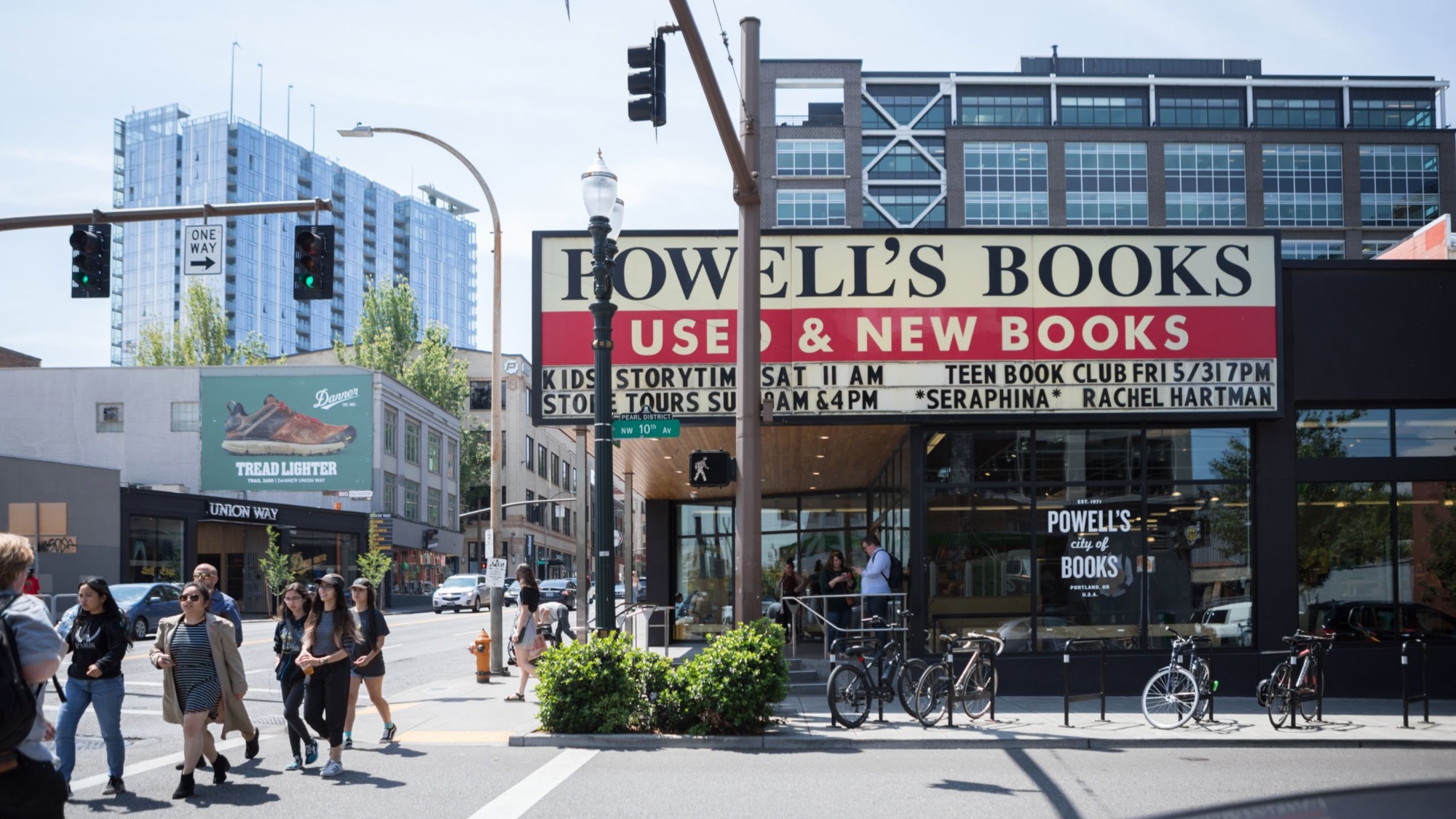 People cross a street in front of Powell's Books, a large bookstore with "Used & New Books" displayed on its sign, in an urban area with modern buildings in the background.