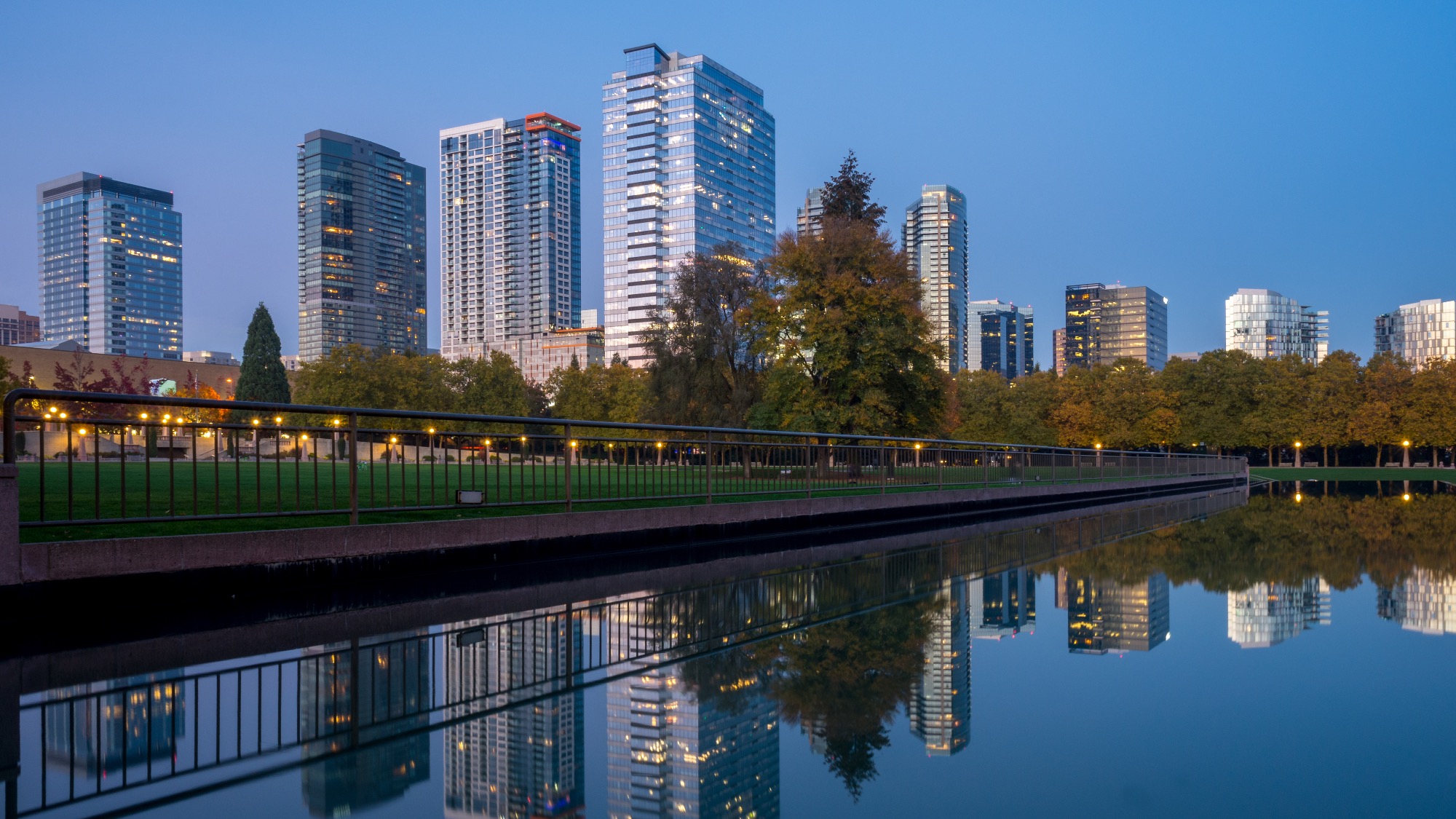 City skyline with modern high-rise buildings and lush trees reflected in a calm body of water at dusk, highlighting Bellevue’s impressive infrastructure.