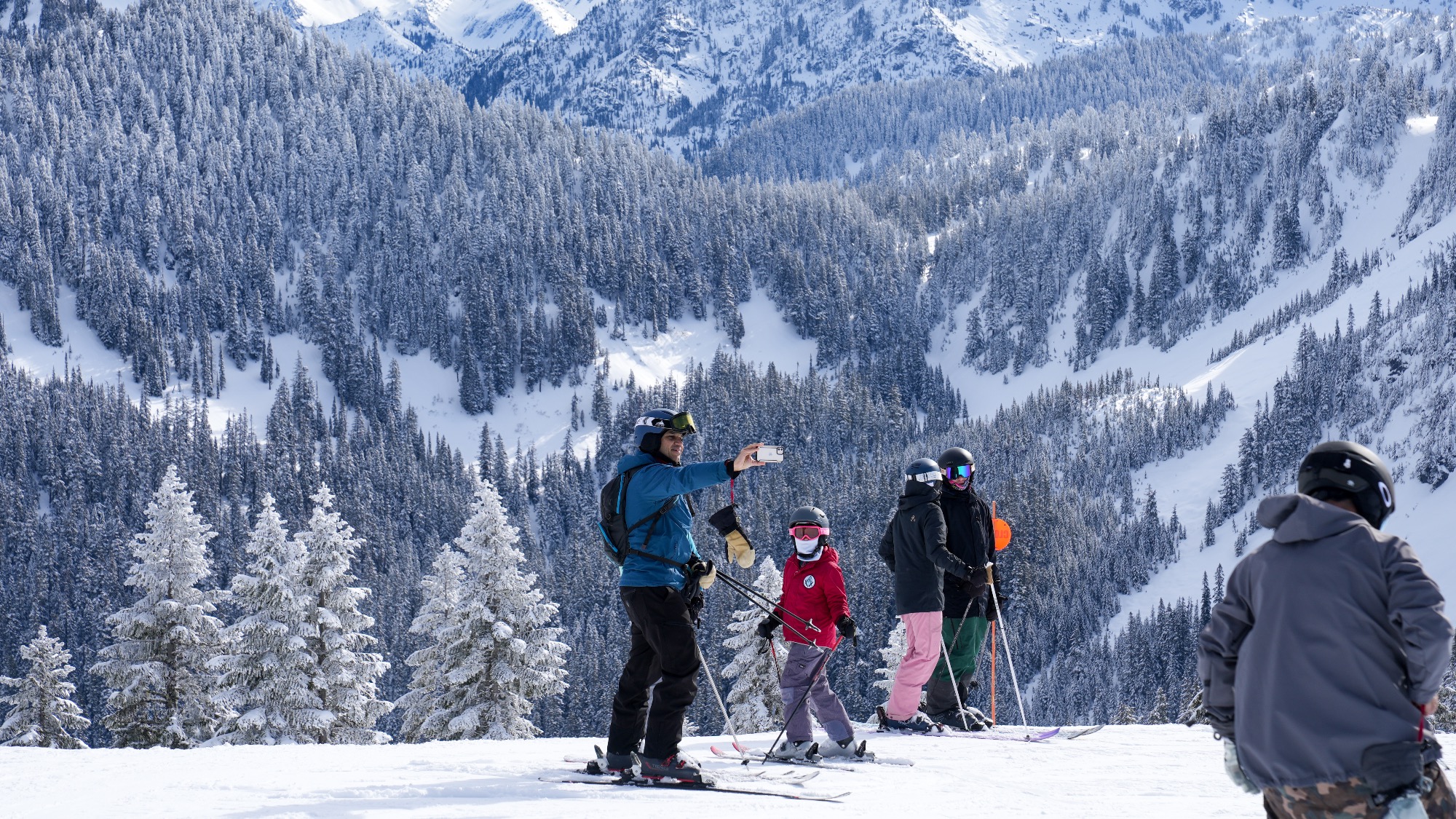 A group of skiers, including adults and children, pause on a snowy slope with mountains and pine trees in the background. One person takes a selfie.