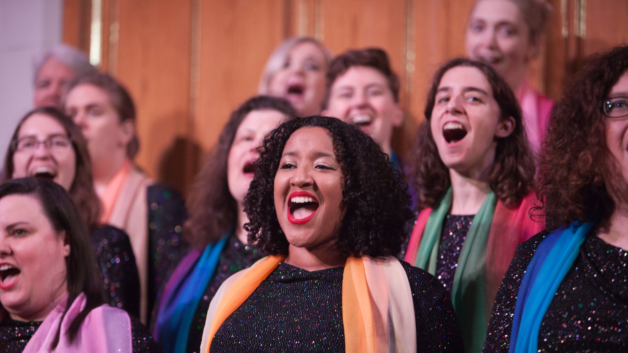 A Seattle Women’s Chorus wearing dark sparkly outfits and colorful scarves sings together in front of a wooden backdrop, sharing women’s stories through powerful harmonies.