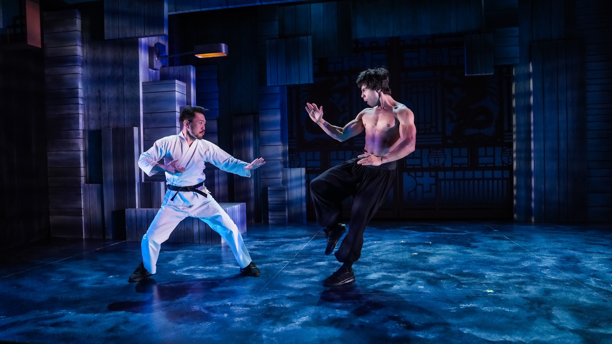 Two men face each other in a martial arts stance on a stage; one wears a white karate uniform, the other is shirtless in black pants. The background features blue-toned set decor.
