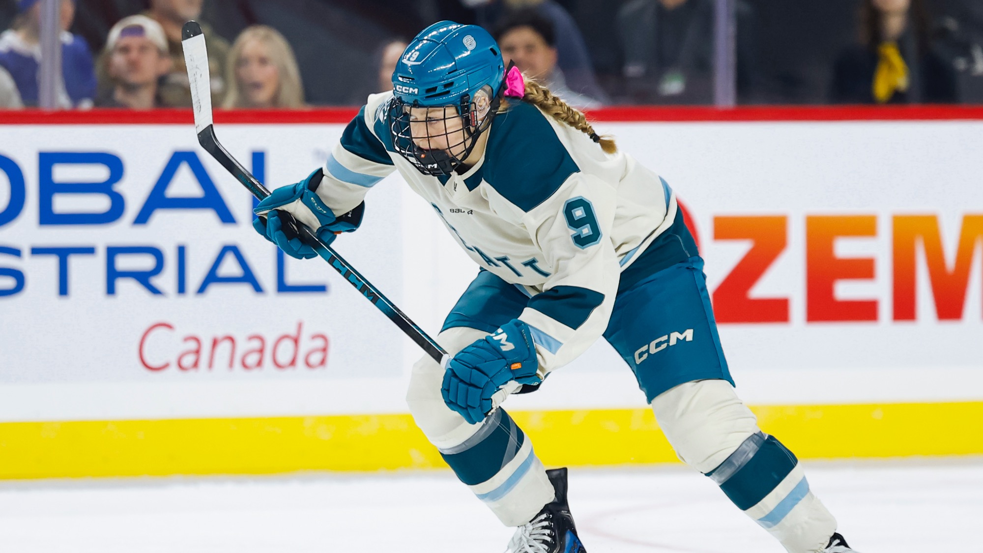 Danielle Serdachny, wearing white and teal gear with the number 9, skates on the ice in a Women's Hockey League game, holding her hockey stick as spectators watch from the background.
