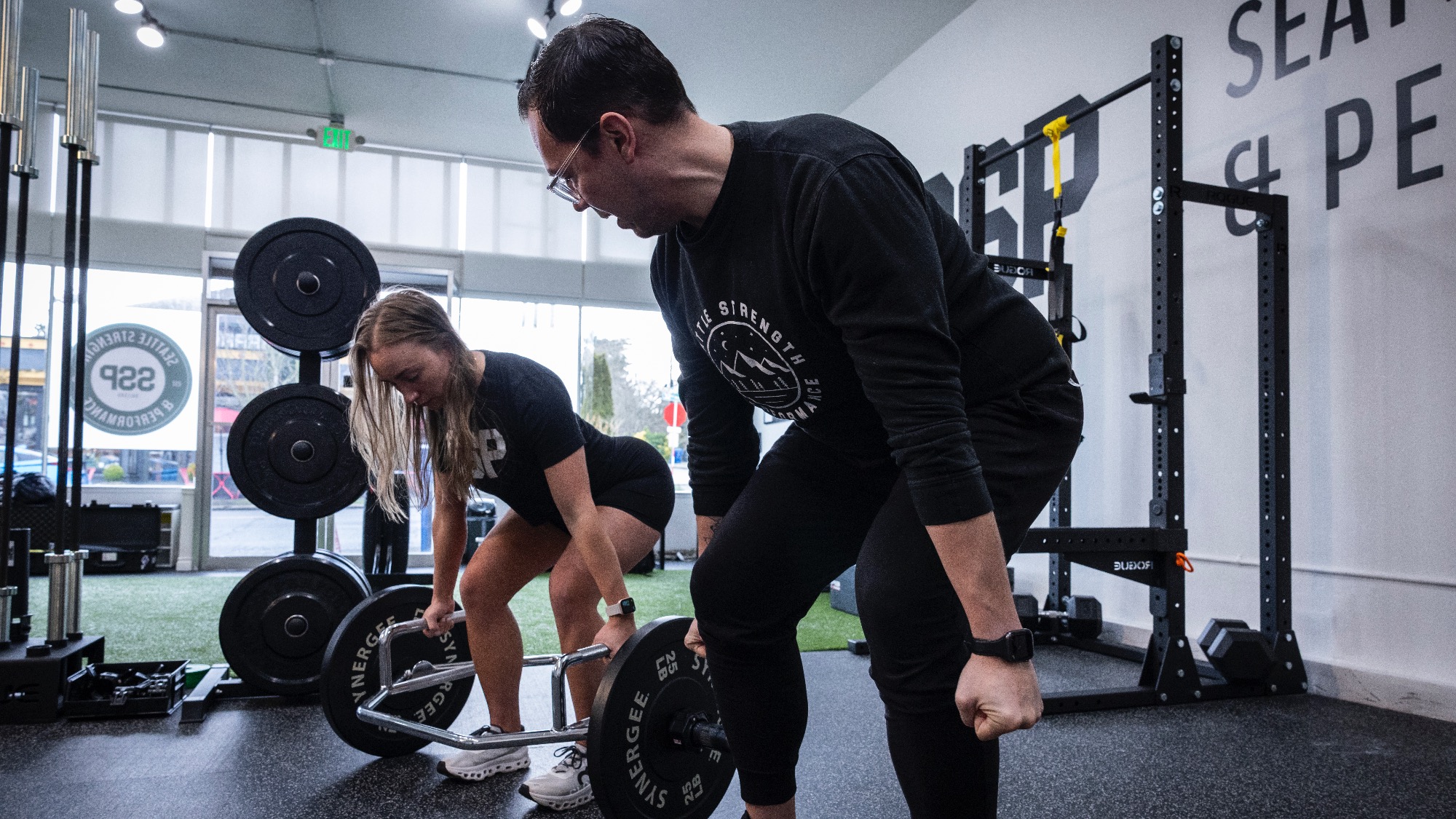 A woman lifts a barbell while a man observes and instructs her inside a gym with various weight equipment.