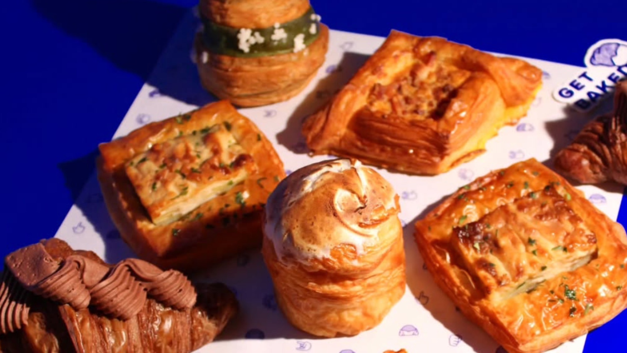 An assortment of pastries, including rectangular, round, and croissant-shaped items, displayed on a branded paper atop a blue surface.