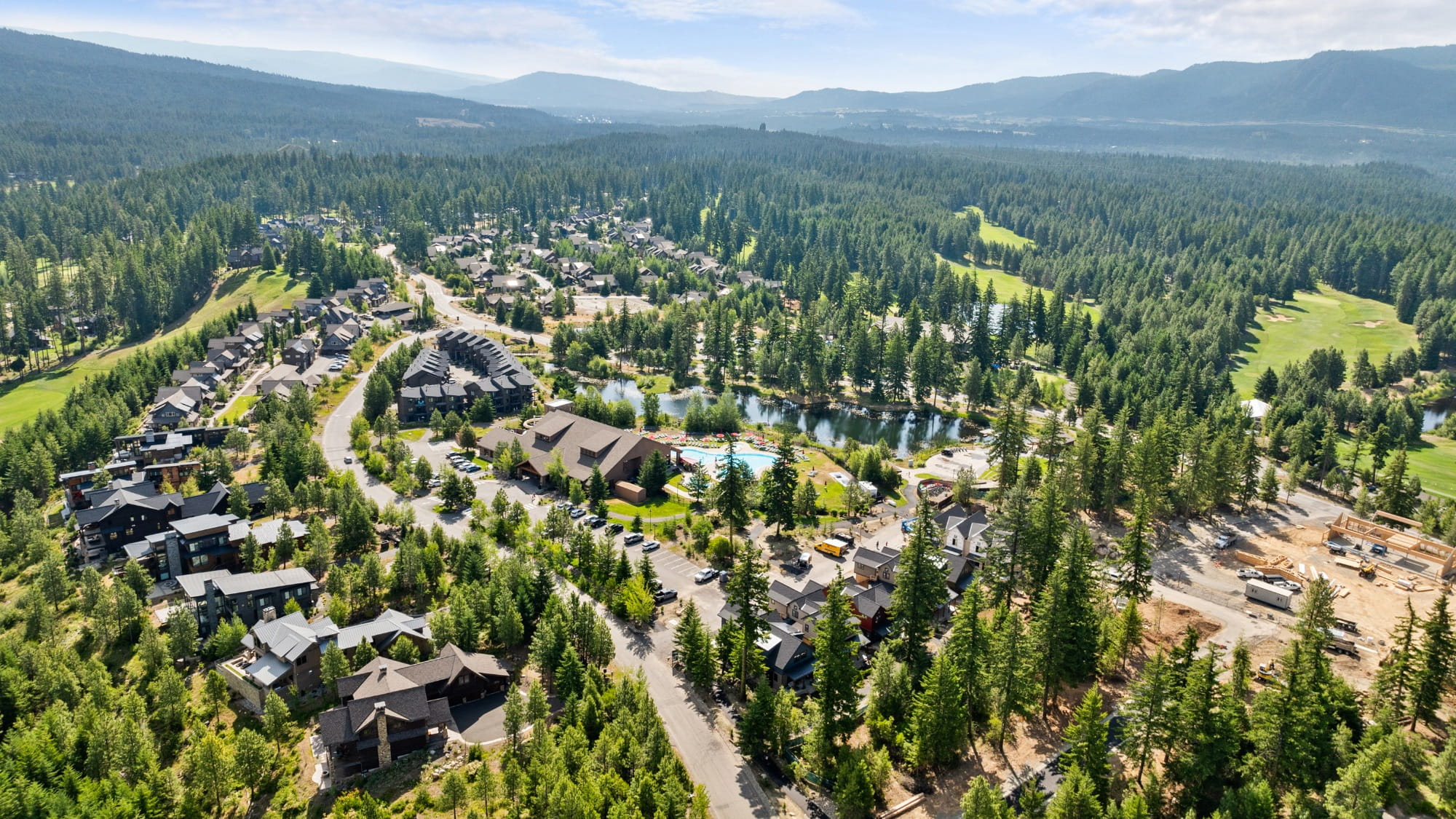 Aerial view of a residential neighborhood in Suncadia, with houses and roads nestled amid dense forest, a pond, and mountains—perfect land to build your dream home.