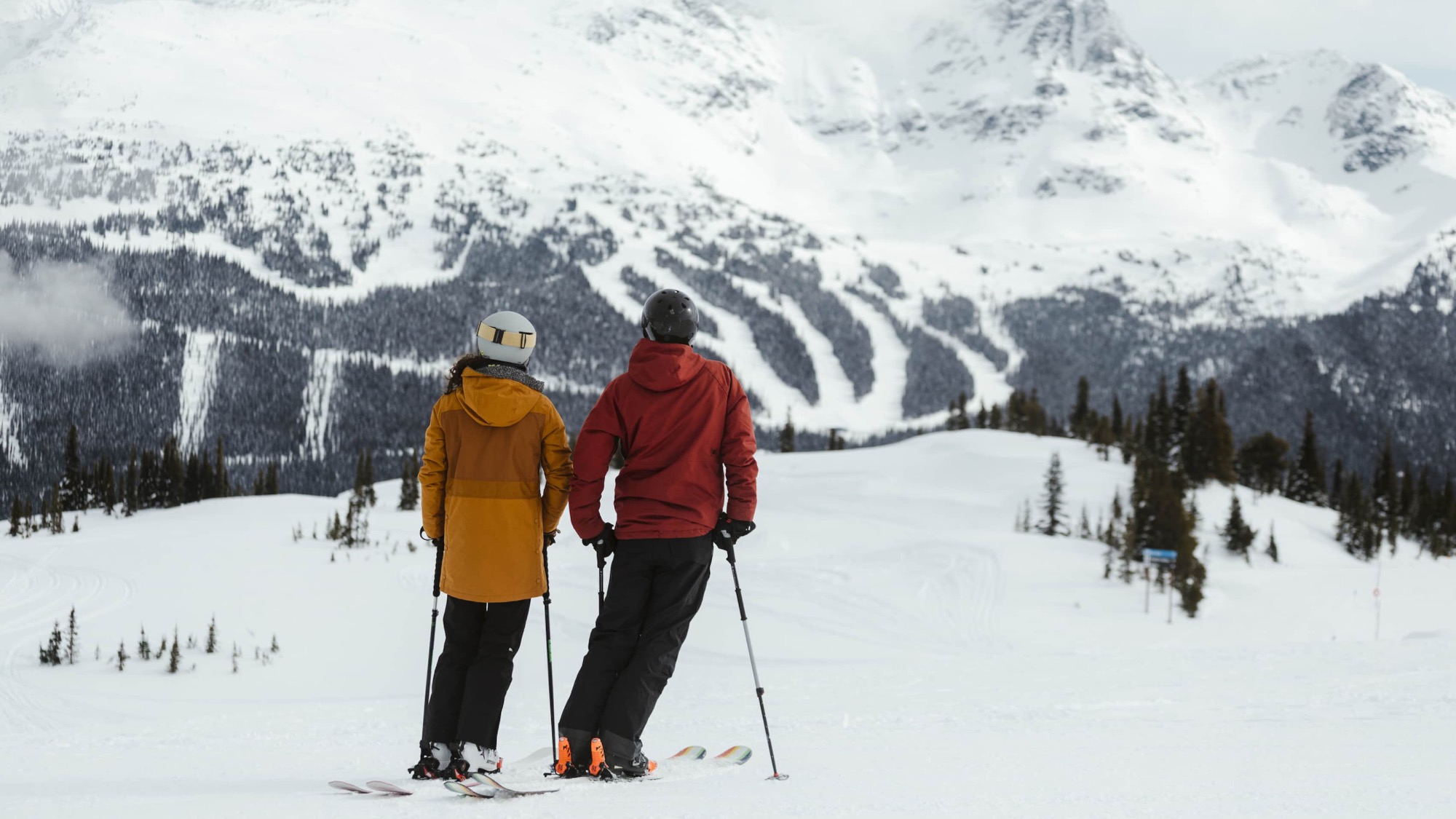 Two people in winter jackets and helmets stand on a snowy slope with skis, taking in the snow-covered mountains—a slow pace for a winter adventure and the perfect time to reset.