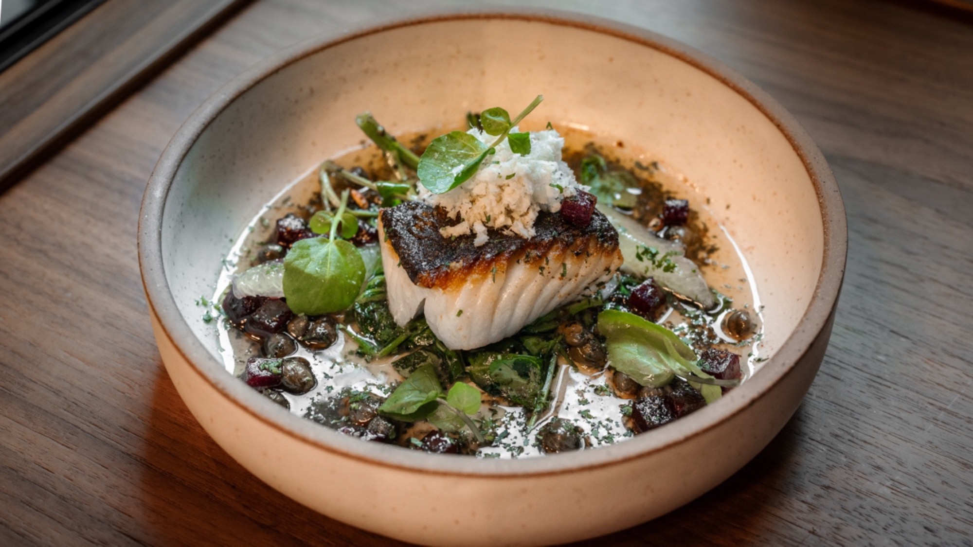 A seared fish fillet garnished with greens and herbs, served in a shallow bowl with a light sauce, capers, and other small vegetable toppings—a vibrant nod to Nordic cuisine.