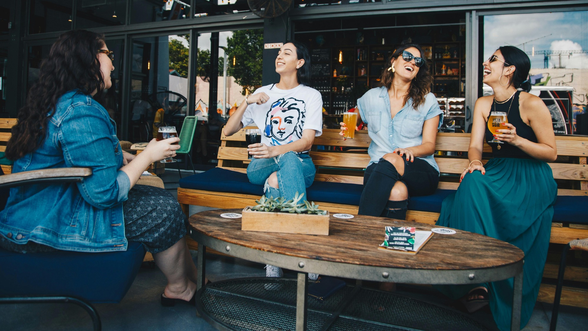 Four women sit on outdoor benches at a bar, holding drinks and laughing together around a table with a small plant centerpiece.