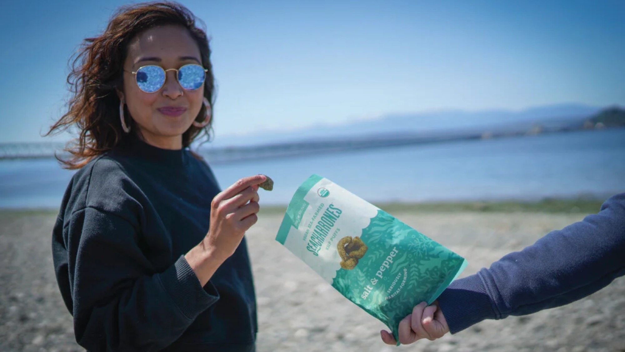A woman on a beach takes a snack from a bag labeled “Sea Salt & Pepper Seaweed” that is being held by another person.