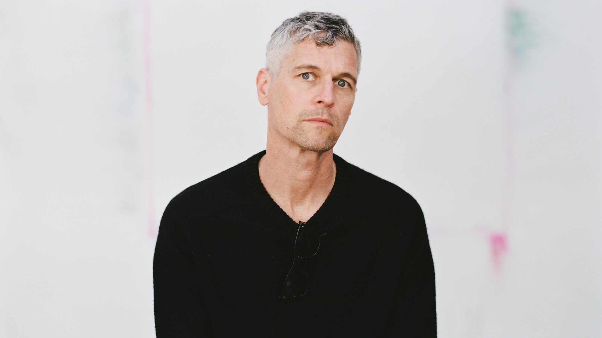 A man with short gray hair wearing a black sweater and glasses sits in front of a plain white background.