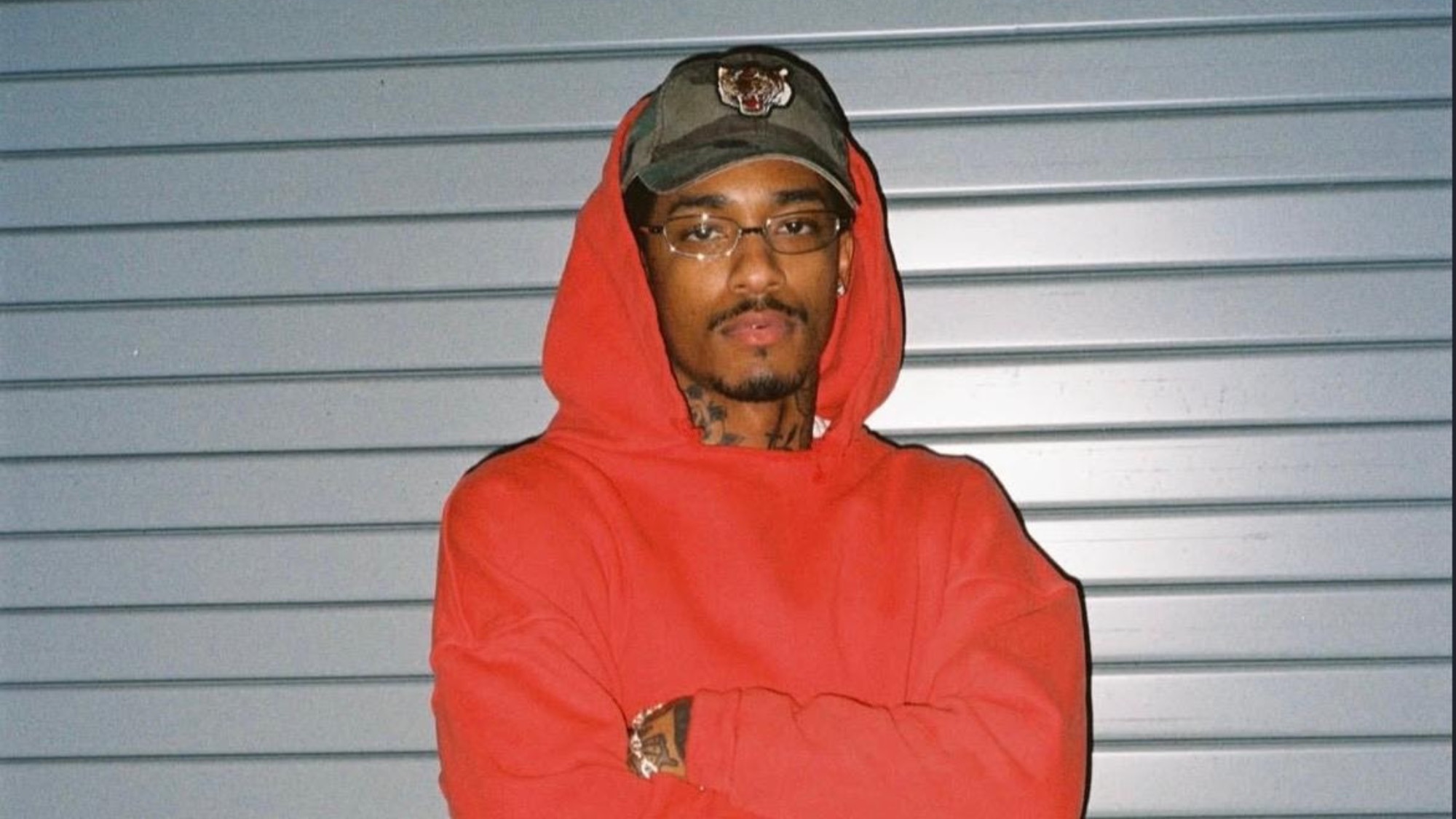 A person wearing a red hoodie, glasses, and a baseball cap stands with arms crossed in front of a metal shutter, exuding the cool confidence of a Slow Burn R&B vibe.