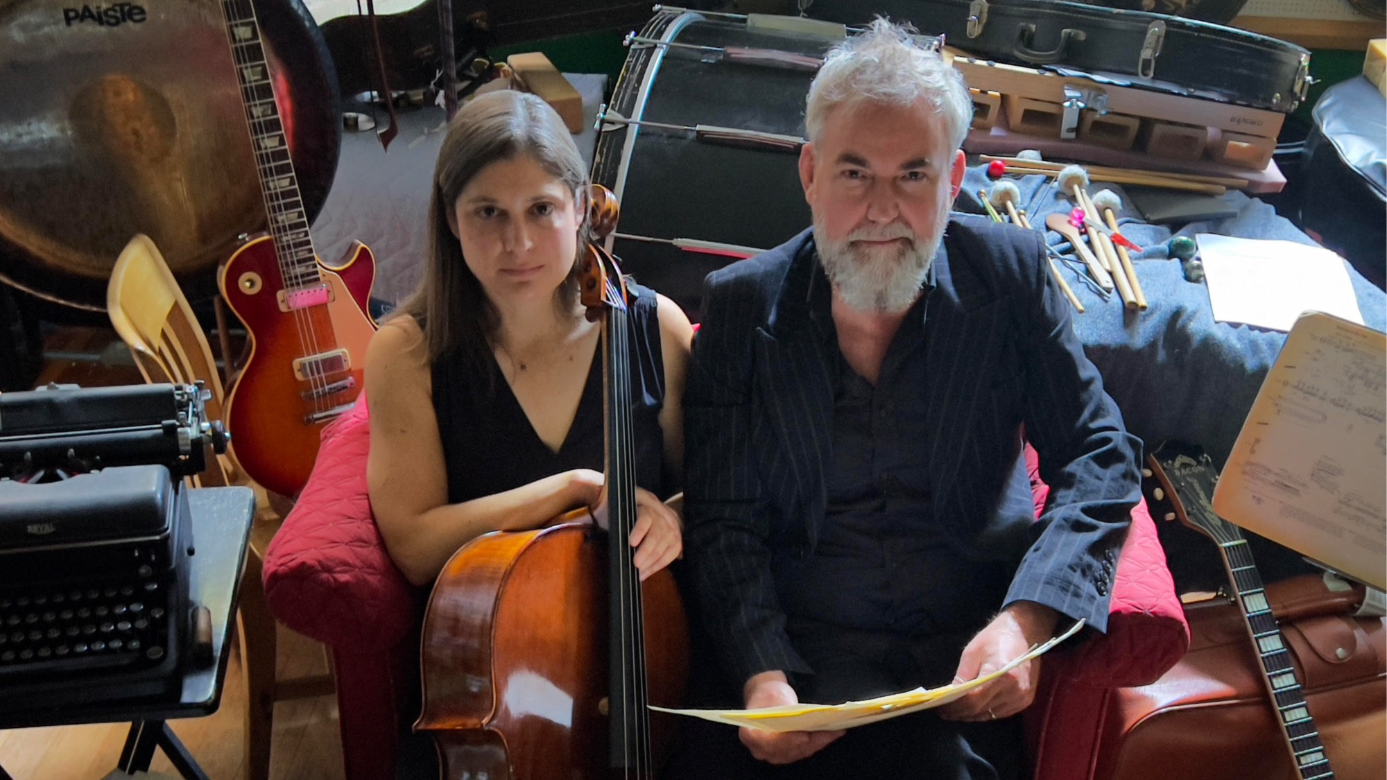 Two people sit on a red chair surrounded by musical instruments, including a cello, guitars, drums, and a typewriter. One holds a cello, the other holds sheet music.
