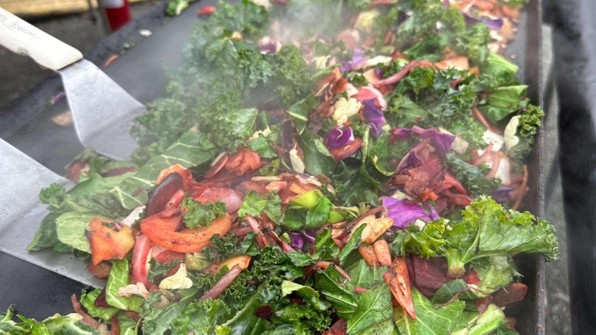A close-up view of assorted colorful vegetables, including kale, carrots, and cabbage, sizzling on a grill with visible steam rising—perfect for creating fresh March dishes full of vibrant food flavors.