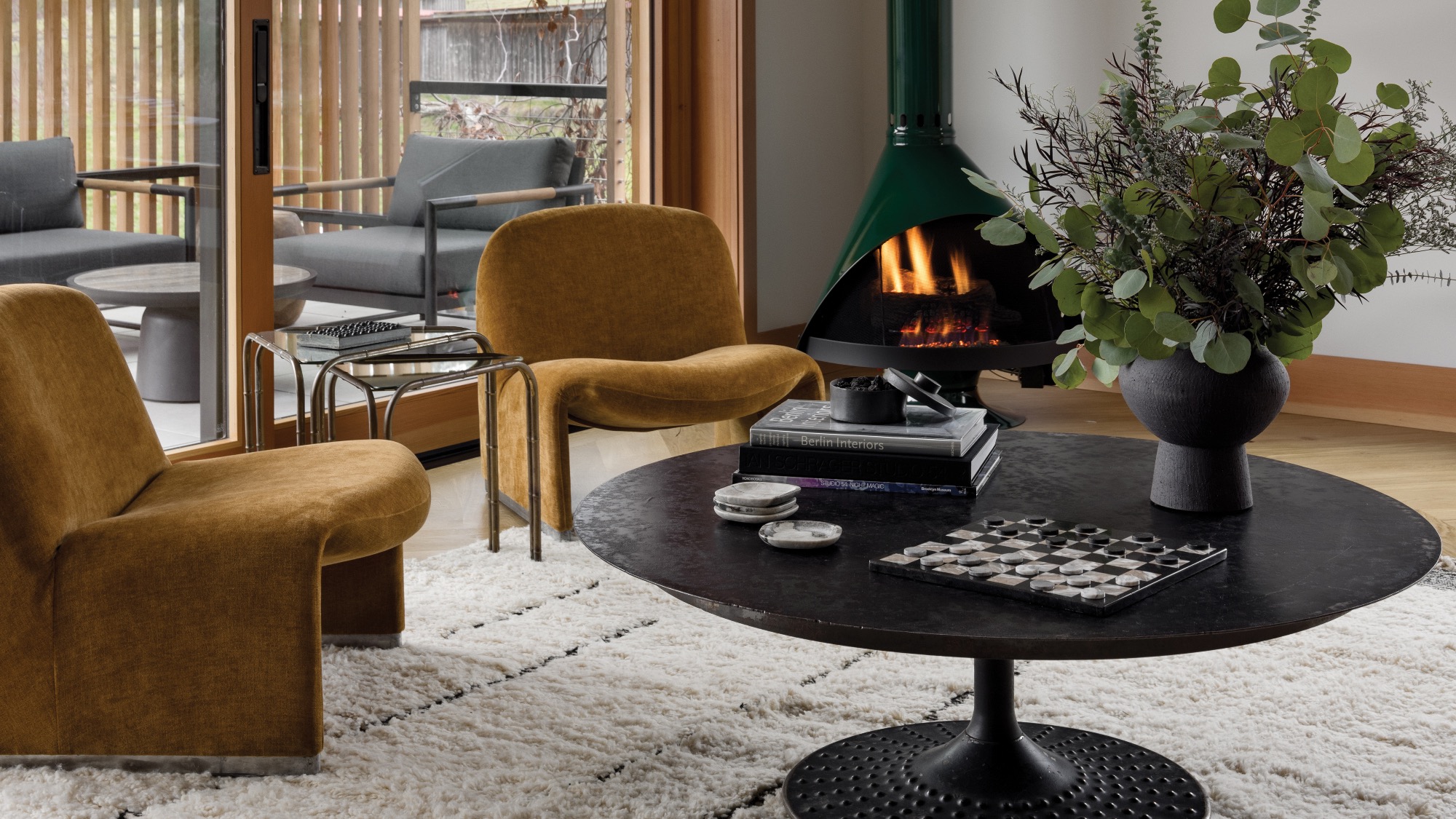 A modern living room with mustard chairs, a round black coffee table, a suspended fireplace with a fire burning, and a large vase with greenery on a white textured rug.