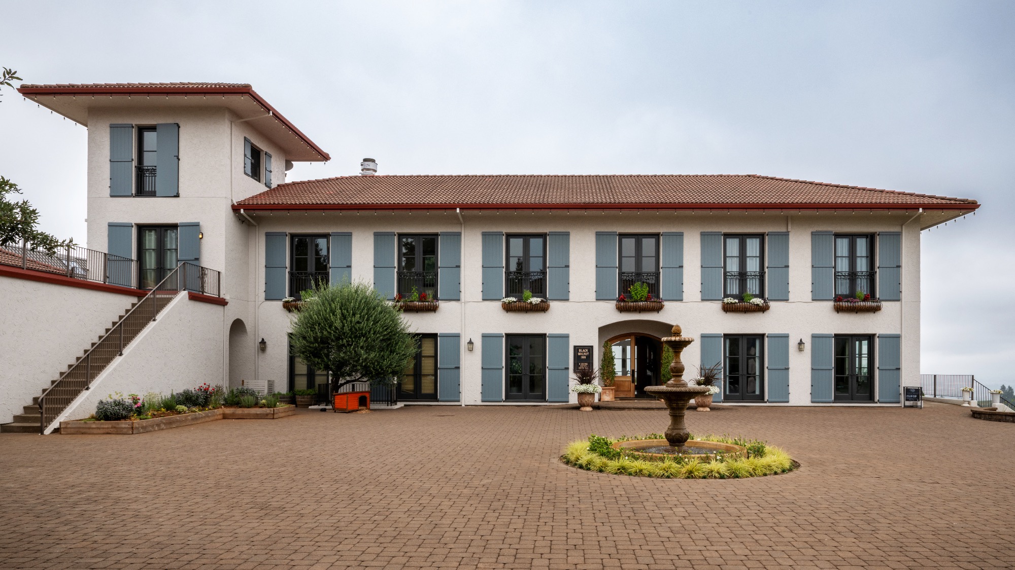 A two-story building with light-colored walls, blue shutters, and a red tile roof, featuring a central fountain in a paved courtyard.