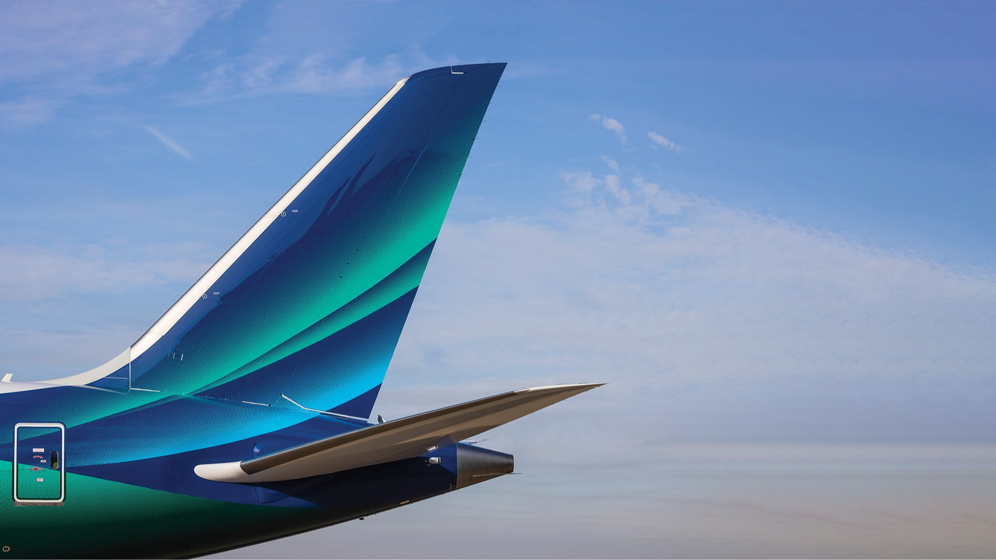 The vertical stabilizer and part of the horizontal stabilizer of an airplane with a blue and green design are shown against a clear sky.