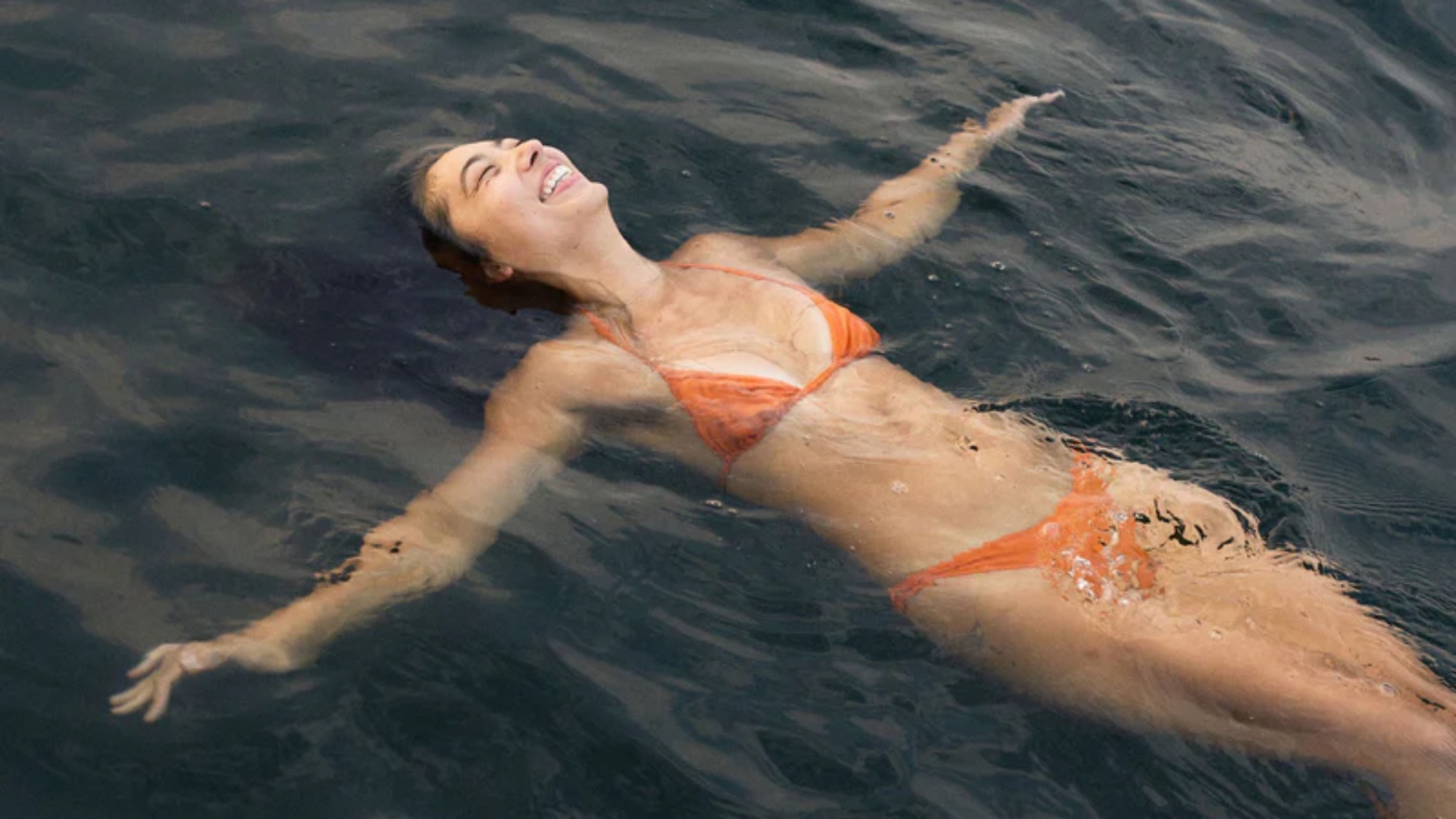 A person in an orange bikini floats on their back in dark water, arms outstretched and face turned upward, appearing relaxed.