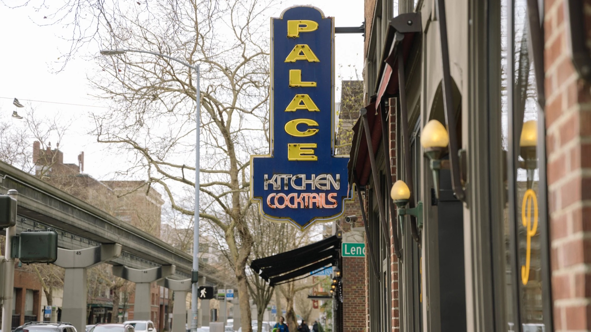 A vertical neon sign reads "Palace Kitchen Cocktails" outside a building on a city street with trees and cars visible in the background.
