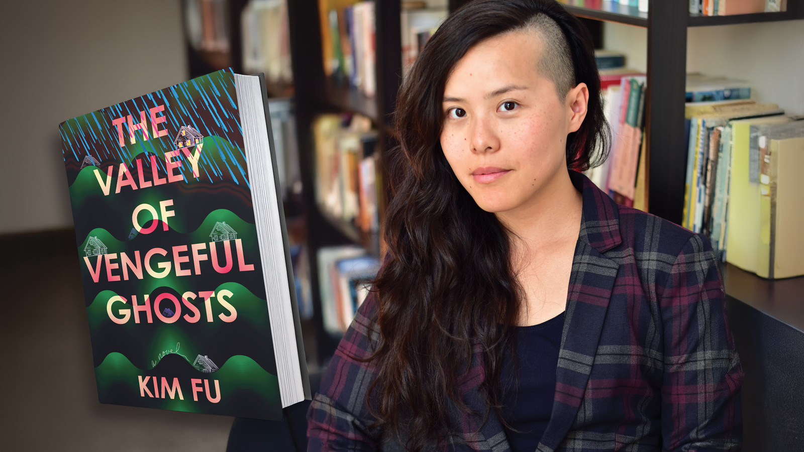A person with long dark hair and an undercut sits in a library next to "The Valley of Vengeful Ghosts" by Kim Fu.