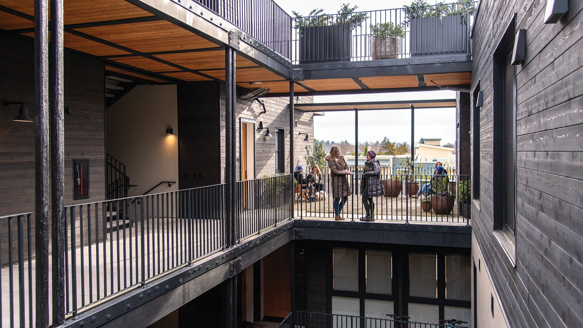 Three people stand and converse on an upper walkway of a modern, open-air building with black railings and wooden ceilings, embodying a Better Together spirit. Potted plants and outdoor seating are visible in the background.