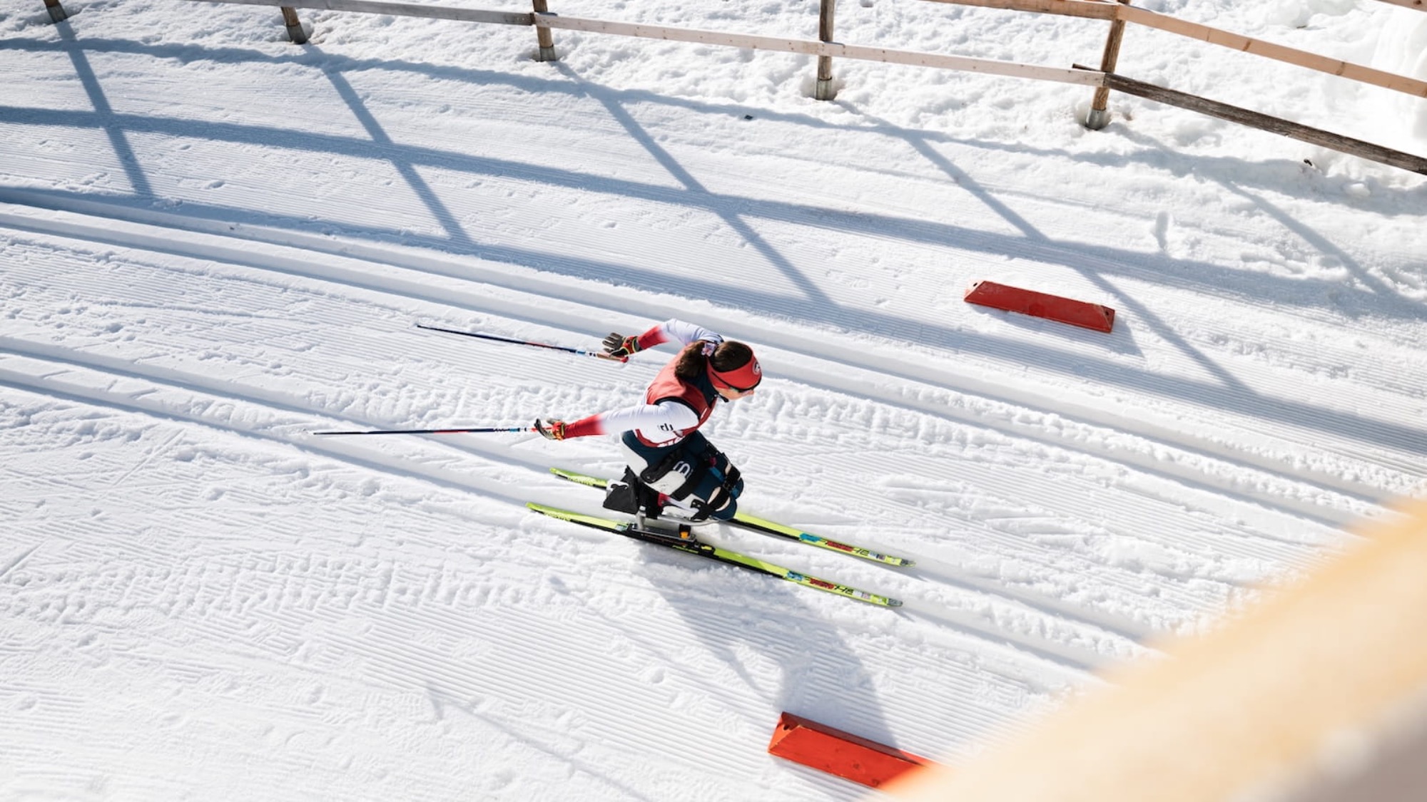 A skier wearing a red and white outfit glides along groomed cross-country ski tracks on a snowy course bordered by a wooden fence.