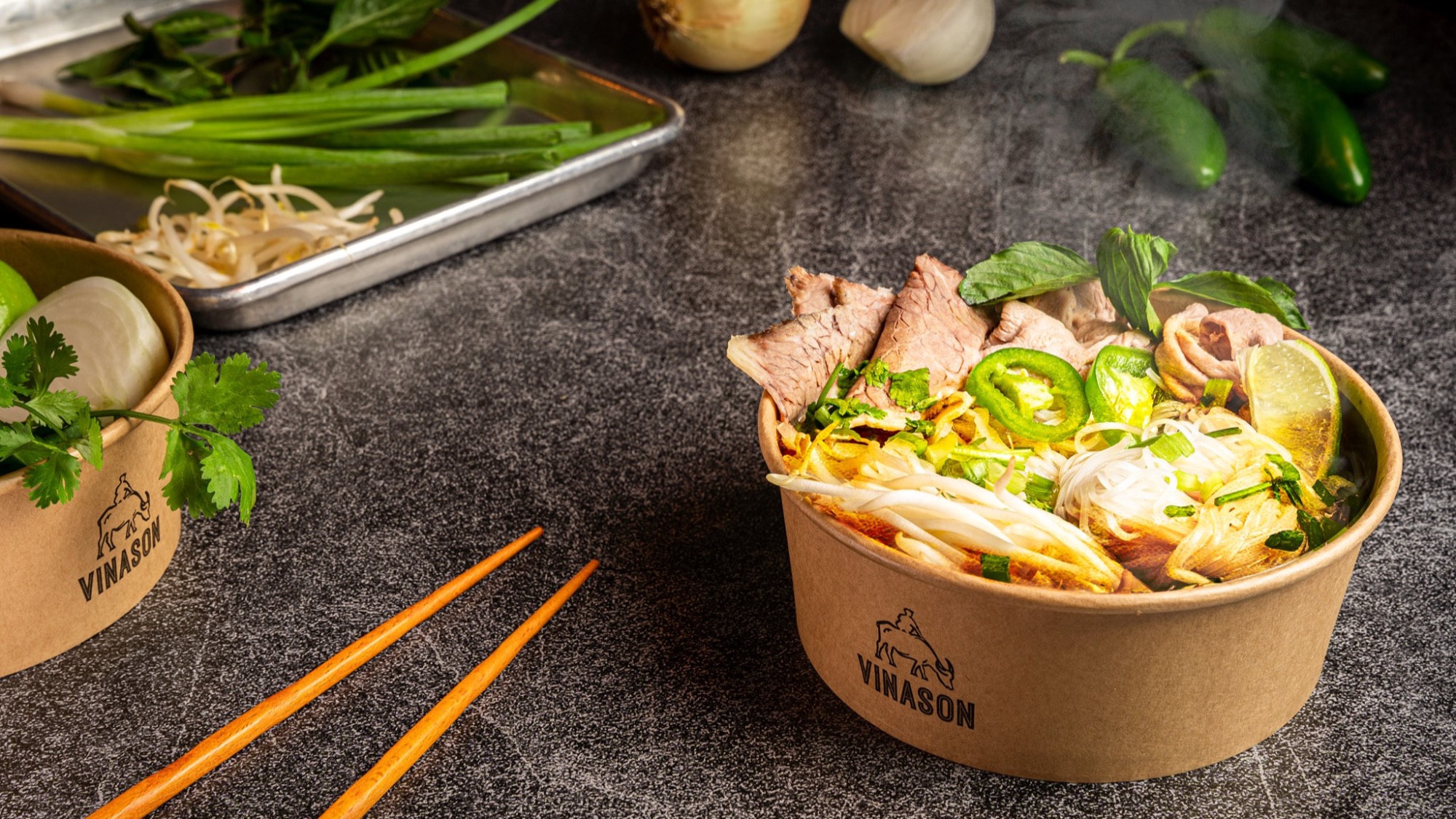 A bowl of Vietnamese pho with sliced beef, noodles, herbs, and lime sits on a counter next to orange chopsticks, fresh vegetables, and another takeout container.