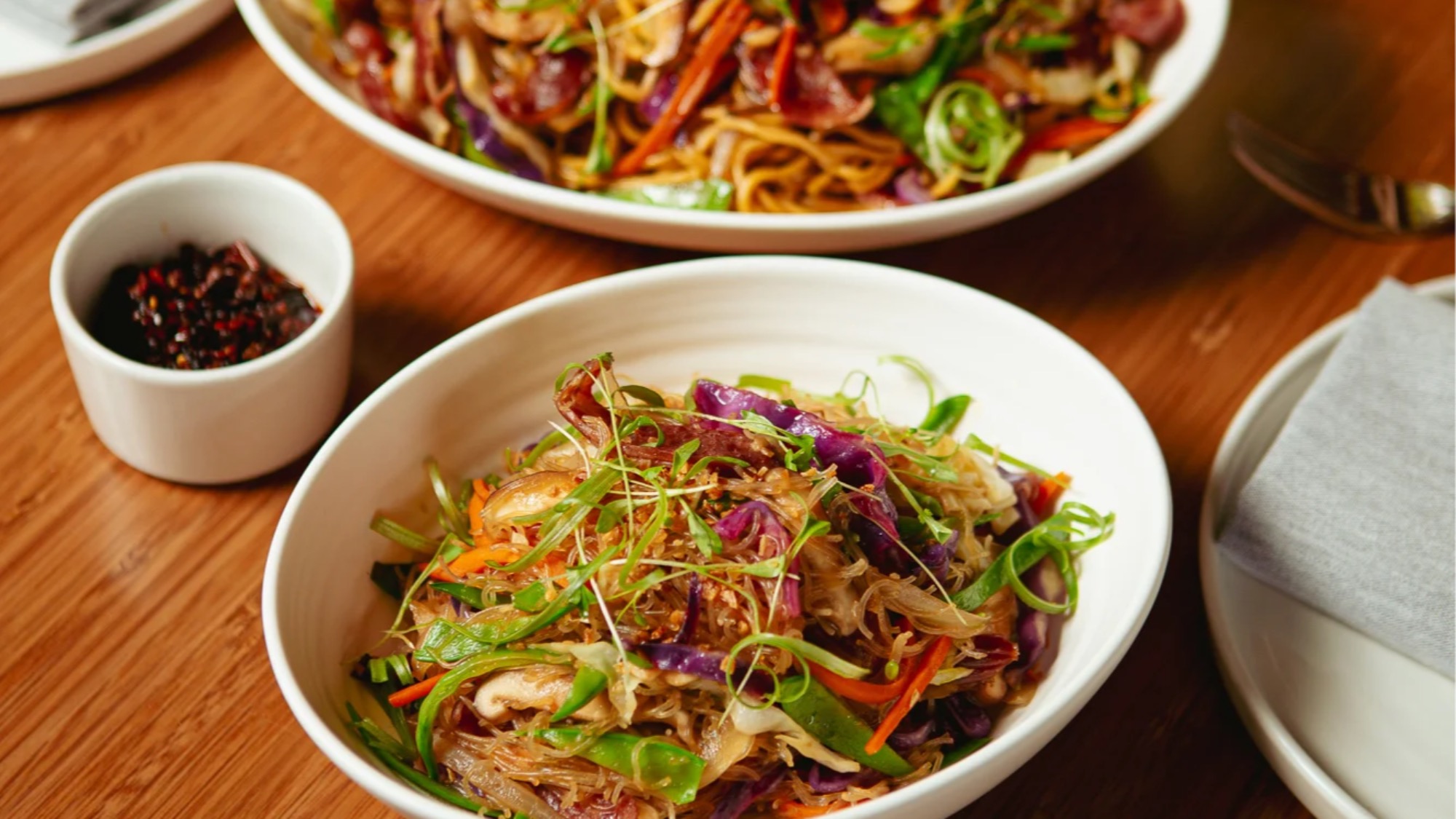 Two bowls of stir-fried noodles with vegetables and greens on a wooden table, accompanied by a small dish of sliced red chili peppers.