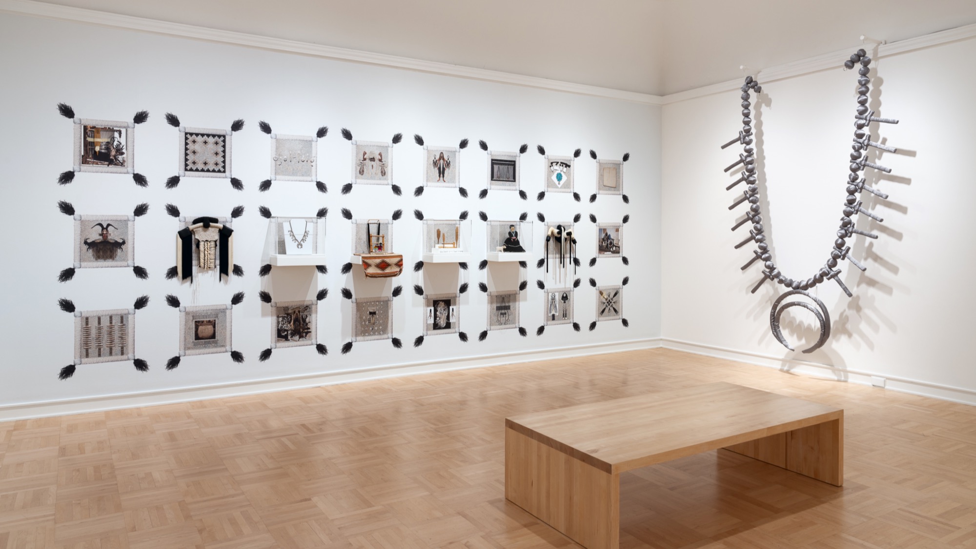 A gallery room with a grid of framed artworks on one wall, a large sculptural necklace on another, and a simple wooden bench on a parquet floor.