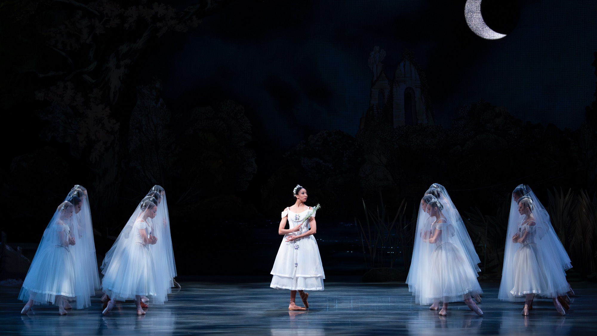 A ballerina in a white costume stands center stage, surrounded by dancers in white dresses and veils, beneath a crescent moon in a dark, forested setting that evokes the elegance of ballet performed at a sculpture park.