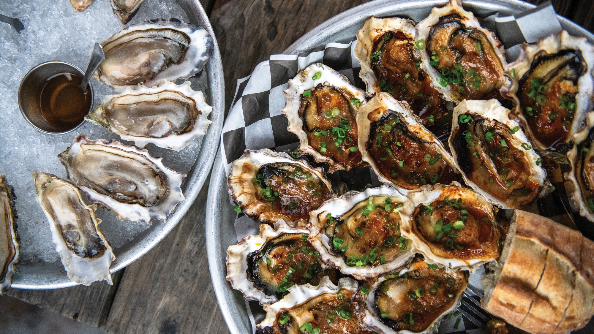 Two trays of oysters: one with raw oysters on ice and sauce, the other with baked oysters topped with herbs and sauce, served with slices of bread on a wooden table.