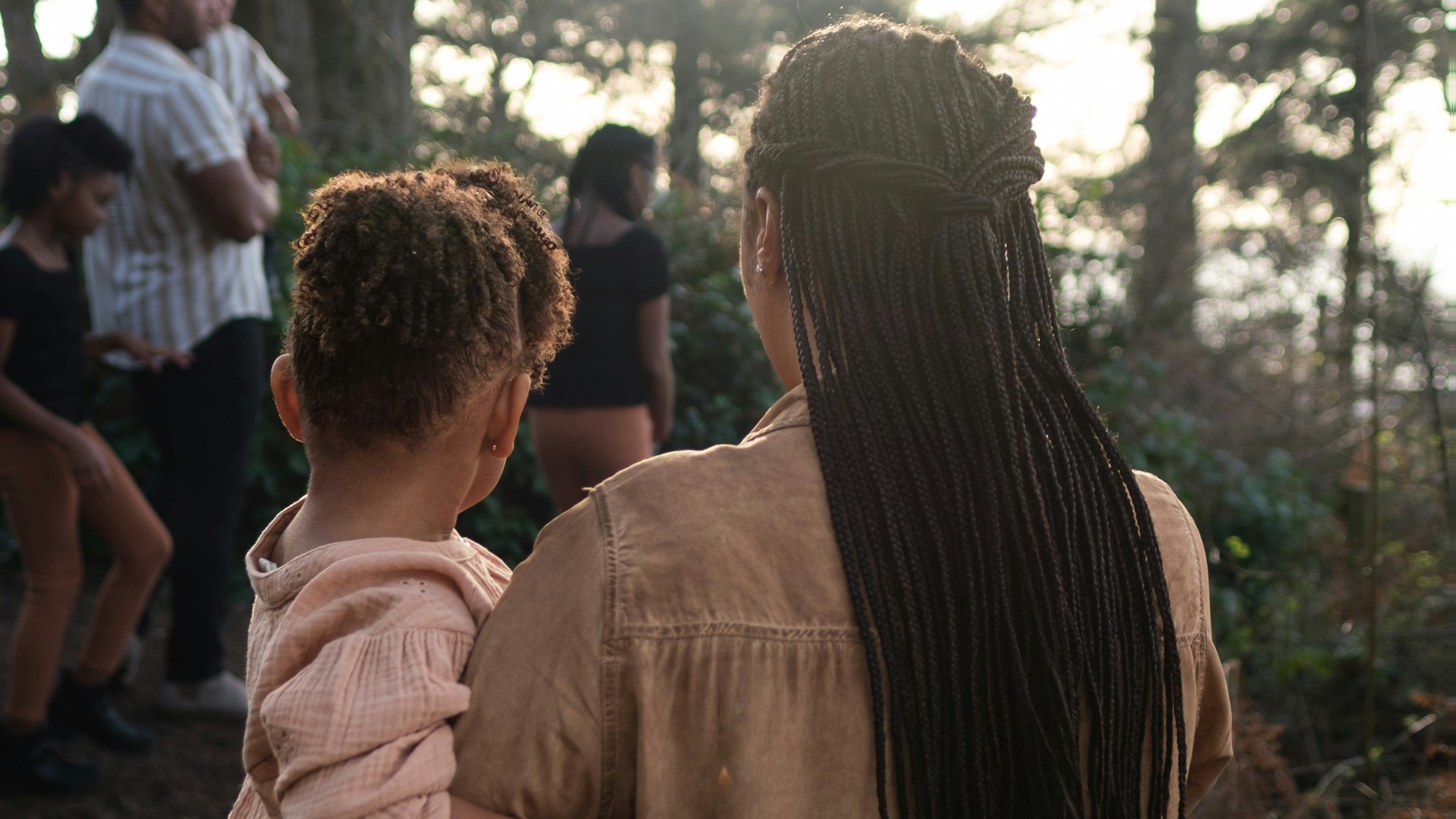 A woman with braids holds a child while standing outdoors in a wooded area with several other people visible in the background.